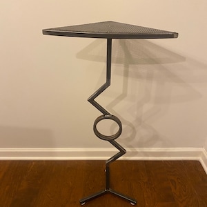 May include: A black metal corner shelf with a triangular top and a geometric base. The shelf has a circular design element in the middle of the base.