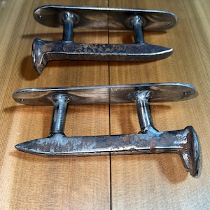 May include: Two antique metal door pulls with a weathered, rustic appearance. Each pull features a long, rectangular base with two vertical supports and a curved handle. The metal has a dark, aged patina, suggesting vintage craftsmanship.