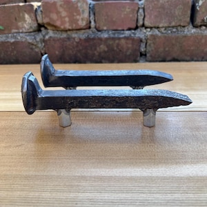 May include: Two weathered, dark metal railroad spike drawer pulls. Each pull has a rectangular head and a tapered, pointed body. The pulls are mounted on small, silver-colored metal supports. The background features a brick wall and a wooden surface.