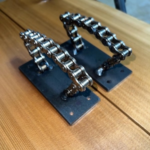 May include: Two industrial-style door pulls made from repurposed bicycle chains. The silver chains are welded to rectangular black metal plates. The pulls are mounted on a light brown wooden surface.