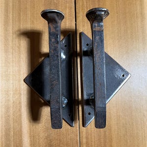 May include: Two rustic, metal door pulls. Each pull features a rectangular handle with a rounded top, attached to a diamond-shaped plate. The hardware is dark gray and appears to be made of iron or steel, set against a light wood background.