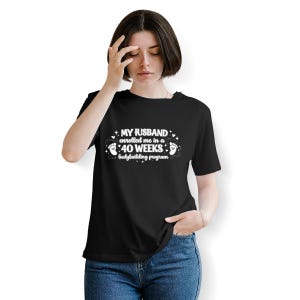 May include: Black t-shirt with white text: "MY HUSBAND enrolled me in a 40 WEEKS bodybuilding program." The text is surrounded by stars and footprints. The shirt is worn by a person.