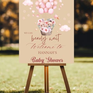 May include: A baby shower welcome sign on a wooden easel. The sign features a hot air balloon with teddy bears, flowers, and balloons. Text reads "We can bearly wait Welcome to Hannah's Baby Shower."