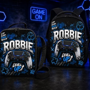 May include: A black backpack and matching lunch bag set featuring a blue and white graphic print of a video game controller, the name ROBBIE, and the text EAT SLEEP GAME REPEAT and LEVEL UP. Both items have a dark theme with neon blue accents.