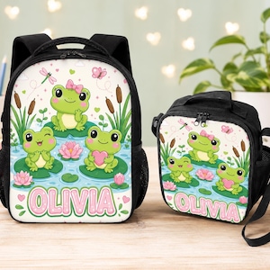 Pode incluir: Uma mochila preta e uma lancheira preta a condizer, ambas com um design estampado de três rãs verdes em folhas de nenúfar, nenúfares cor-de-rosa, taboas e libélulas. O nome OLIVIA está impresso em letras cor-de-rosa grandes na parte inferior.