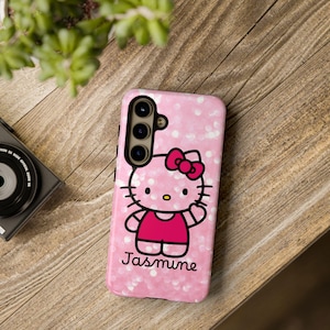 Puede incluir: Funda de teléfono rosa con un diseño de Hello Kitty, lazo y atuendo rojos. La funda tiene un fondo brillante y el nombre "Jasmine" en cursiva. La funda está sobre una superficie de madera.