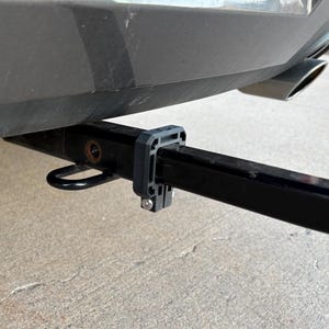 May include: A black trailer hitch receiver attached to the rear of a vehicle. The hitch includes a black receiver tube, a safety chain loop, and a black plastic bracket. The vehicle's bumper is gray, and the exhaust pipe is visible.