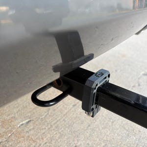 Puede incluir: Un enganche de remolque negro con un lazo negro y un soporte gris. El enganche est&aacute; fijado a la parte inferior de un veh&iacute;culo plateado. El enganche est&aacute; dise&ntilde;ado para remolcar.