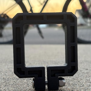 Puede incluir: Accesorio de bicicleta negro, de forma cuadrada, con una abertura rectangular. Cuenta con una abrazadera con tornillo y tuerca para la fijaci&oacute;n. En el fondo se ve una bicicleta y un atardecer borroso.
