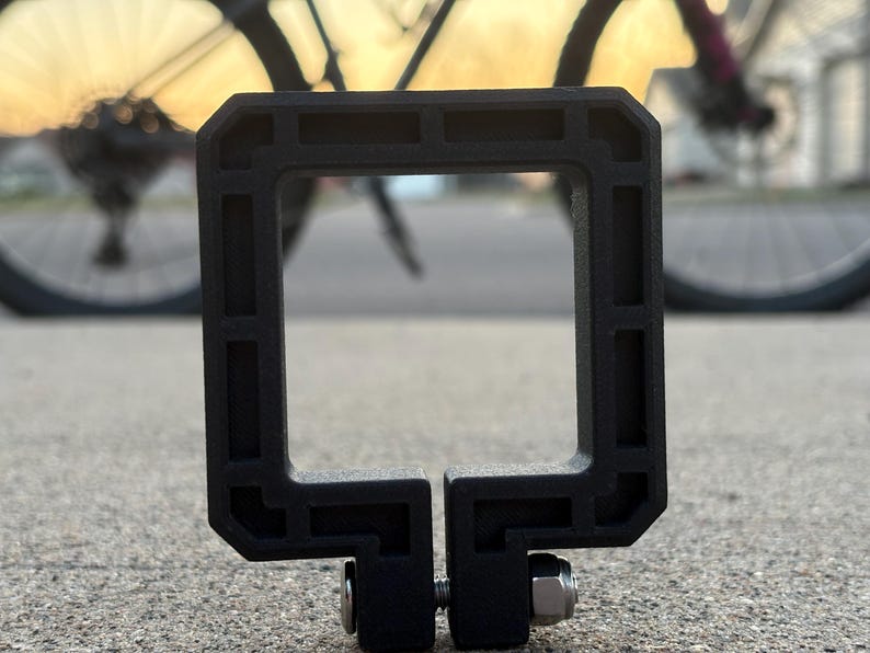 Puede incluir: Accesorio de bicicleta negro, de forma cuadrada, con una abertura rectangular. Cuenta con una abrazadera con tornillo y tuerca para la fijaci&oacute;n. En el fondo se ve una bicicleta y un atardecer borroso.