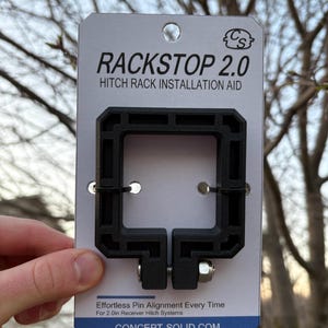 Puede incluir: Ayuda de instalaci&oacute;n de portaequipajes de enganche Rackstop 2.0 negro. El producto est&aacute; sostenido por una mano. El producto es un cuadrado negro con un perno y el texto "Effortless Pin Alignment Every Time" y "CONCEPT-SOLID.COM".