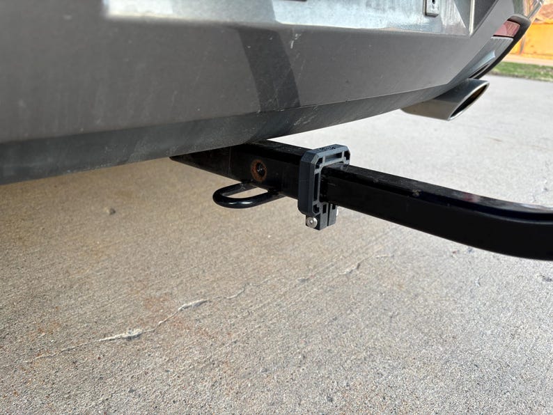 May include: A black trailer hitch receiver attached to the rear of a vehicle. The hitch includes a black receiver tube, a safety chain loop, and a black plastic bracket. The vehicle's bumper is gray, and the exhaust pipe is visible.