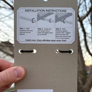 Puede incluir: Una tarjeta de embalaje de producto de cart&oacute;n marr&oacute;n con instrucciones de instalaci&oacute;n para un enganche RackStop. La tarjeta tiene un agujero en la parte superior y dos bandas el&aacute;sticas negras. El texto incluye los pasos de instalaci&oacute;n y una advertencia de peligro de asfixia.