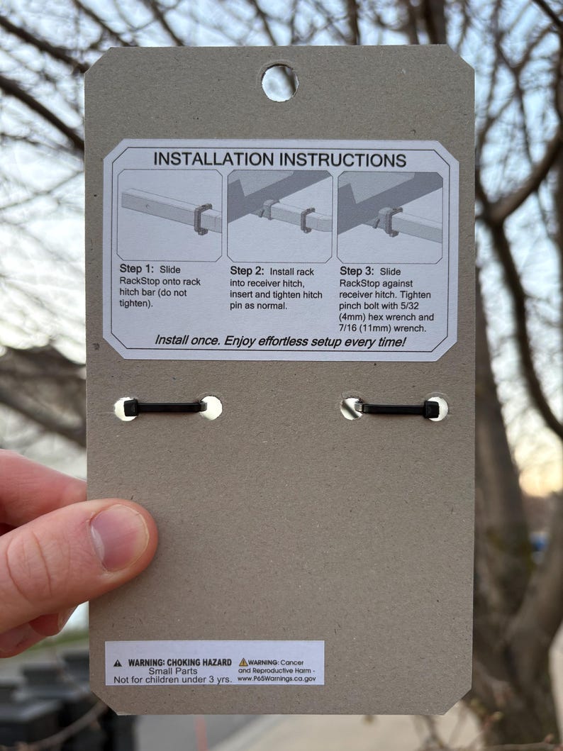 Puede incluir: Una tarjeta de embalaje de producto de cart&oacute;n marr&oacute;n con instrucciones de instalaci&oacute;n para un enganche RackStop. La tarjeta tiene un agujero en la parte superior y dos bandas el&aacute;sticas negras. El texto incluye los pasos de instalaci&oacute;n y una advertencia de peligro de asfixia.