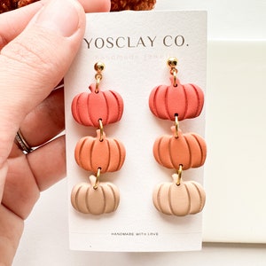Può includere: Un paio di orecchini pendenti con tre ciondoli a forma di zucca impilati in tonalità di arancione, pesca e crema. Gli orecchini sono fatti a mano e hanno elementi dorati. La carta recita "YOSCLAY CO. gioielli fatti a mano" e "FATTO A MANO CON AMORE".