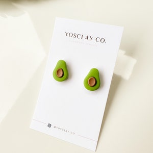 May include: A pair of avocado-shaped earrings made of green and brown polymer clay. The earrings are on a white card with the text "YOSCLAY CO. Handmade Jewelry" and "@YOSCLAY.CO".