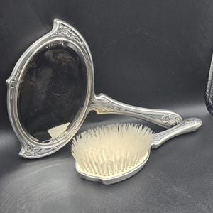 May include: A silver-toned vanity set featuring a hand mirror and a brush. The mirror has an oval shape with decorative floral detailing around the frame. The brush has a matching handle and light-colored bristles.