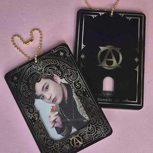 May include: Two black card-shaped photo holders with gold accents and a gold chain. The front of one holder features a photo of a man holding a red flower. The back of the other holder has the text "Blouseide Art" and a gold letter "A".