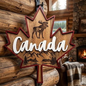 May include: Wooden sign shaped like a maple leaf, with a red border and the word "Canada" in white. It features a moose with a heart and a mountain and forest scene. The sign is mounted on a wooden wall.