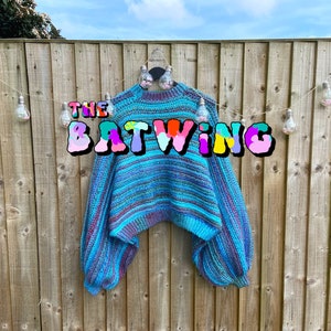 May include: A blue, purple, and teal batwing sweater hangs against a wooden fence. The sweater has a striped pattern and long, voluminous sleeves. The text "THE BATWING" is displayed across the sweater in colorful bubble letters.