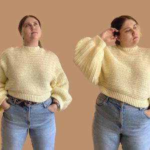May include: A light yellow crochet sweater with a crew neck and long, voluminous sleeves. The sweater is paired with light blue jeans and a patterned belt. The model is wearing a watch. The background is a solid tan color.