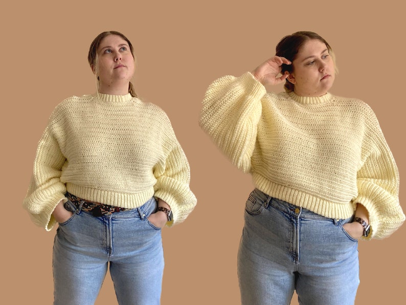 May include: A light yellow crochet sweater with a crew neck and long, voluminous sleeves. The sweater is paired with light blue jeans and a patterned belt. The model is wearing a watch. The background is a solid tan color.