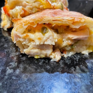 Puede incluir: Un primer plano de un pastel dorado relleno de pollo, queso y verduras. El pastel está cortado por la mitad, revelando el relleno.