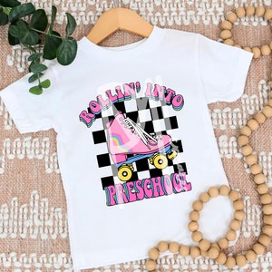 May include: White t-shirt with a pink roller skate graphic and the text "Rollin' into Preschool" in rainbow colors on a black and white checkered background.