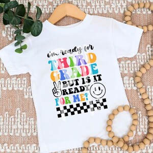 Op de afbeelding: Wit T-shirt met de tekst "I'm ready for third grade but is it ready for me?" in kleurrijke letters met een vredesteken en een smiley.