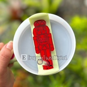 May include: A red nutcracker figurine with a gold crown and a white background. The figurine is on a white lid with a straw sticking out of the top.