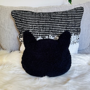 May include: A black plush cat-shaped pillow with large ears sits on a white fluffy surface. The pillow is in front of a black and white patterned pillow.