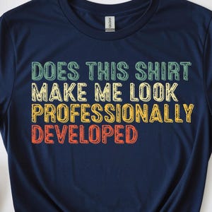 May include: A navy blue short-sleeved t-shirt featuring the text "DOES THIS SHIRT MAKE ME LOOK PROFESSIONALLY DEVELOPED" printed in stacked lines of green, yellow, and orange retro-style block lettering across the center of the chest.