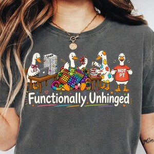 Könnte beinhalten: Dunkelgraues T-Shirt mit einer Grafik von Cartoon-Gänsen, die stricken und einen Laptop benutzen. Der Text "Functionally Unhinged" ist darunter gedruckt.