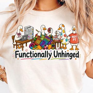 Könnte beinhalten: Elfenbeinfarbenes T-Shirt mit einem skurrilen Design von Cartoon-Enten, die sich mit verschiedenen Aktivitäten wie Stricken und Arbeiten an einem Laptop beschäftigen. Der Satz "Functionally Unhinged" ist unter den Enten in bunten Buchstaben aufgedruckt. Das Shirt hat kurze Ärmel.