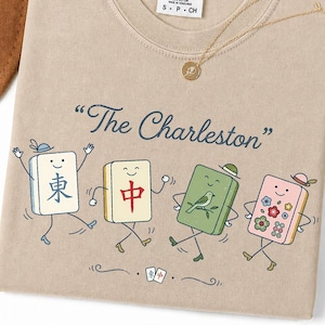 May include: Tan Comfort Colors t-shirt with the text "The Charleston" and dancing mahjong tiles. The tiles are in various colors and feature whimsical designs. A gold necklace with a pendant is also visible.
