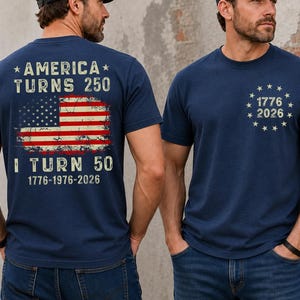 Op de afbeelding: Marineblauwe T-shirts met patriottische ontwerpen. E&eacute;n shirt heeft een versleten Amerikaanse vlag en de tekst "AMERICA TURNS 250" en "I TURN 50 1776-1976-2026." Het andere shirt heeft "1776 2026" omringd door sterren.