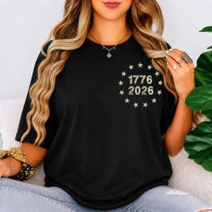 Op de afbeelding: Zwart T-shirt met de tekst "1776 2026" omringd door sterren. Het ontwerp heeft een vintage uitstraling. Het shirt heeft korte mouwen en een ronde hals.