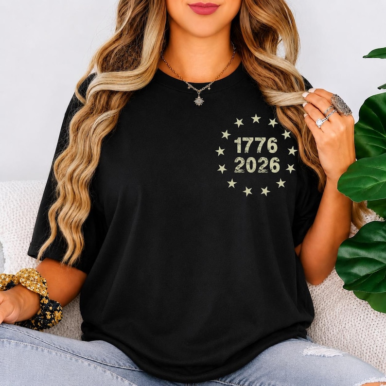Op de afbeelding: Zwart T-shirt met de tekst "1776 2026" omringd door sterren. Het ontwerp heeft een vintage uitstraling. Het shirt heeft korte mouwen en een ronde hals.