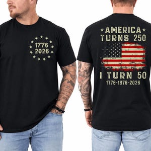 Op de afbeelding: Zwart T-shirt met de tekst "1776 2026" omringd door sterren op de voorkant. De achterkant van het shirt heeft een versleten Amerikaans vlagontwerp met de tekst "AMERICA TURNS 250" en "I TURN 50" met de data 1776-1976-2026.