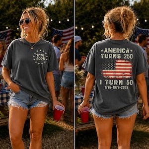 Op de afbeelding: Donkergrijs t-shirt met de tekst "1776 2026" op de voorkant en "America Turns 250, I Turn 50" met een Amerikaanse vlag op de achterkant. Het shirt wordt gedragen met een spijkerbroekshort.