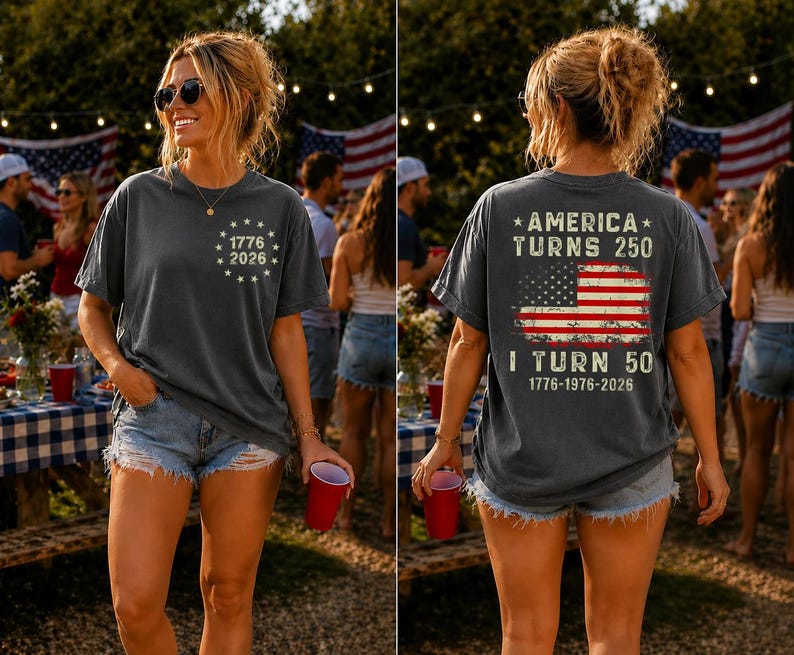 Op de afbeelding: Donkergrijs t-shirt met de tekst "1776 2026" op de voorkant en "America Turns 250, I Turn 50" met een Amerikaanse vlag op de achterkant. Het shirt wordt gedragen met een spijkerbroekshort.