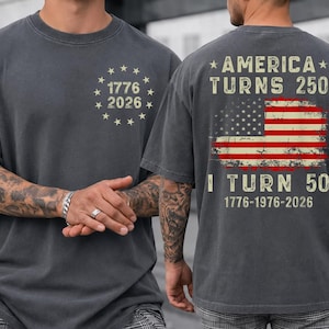 Op de afbeelding: Donkergrijs t-shirt met de tekst "1776 2026" in een cirkel van sterren op de voorkant. De achterkant van het shirt heeft een Amerikaanse vlag en de tekst "America Turns 250, I Turn 50, 1776-1976-2026."