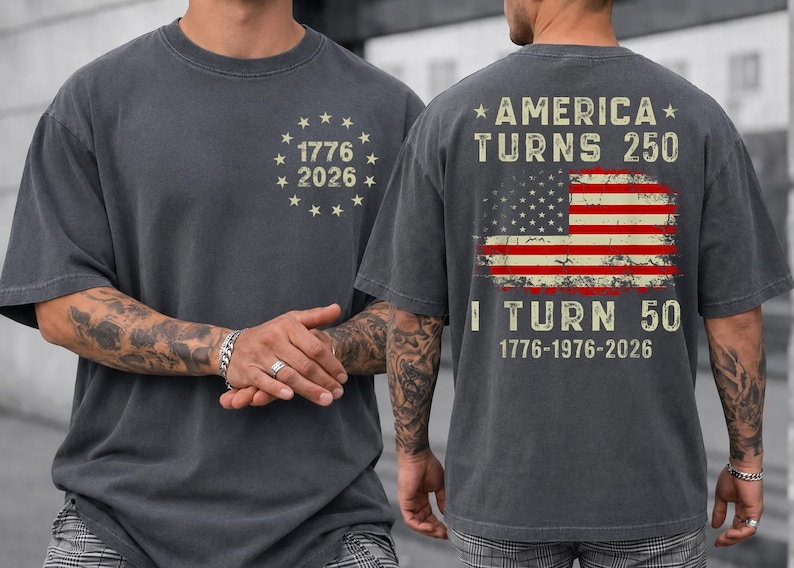 Op de afbeelding: Donkergrijs t-shirt met de tekst "1776 2026" in een cirkel van sterren op de voorkant. De achterkant van het shirt heeft een Amerikaanse vlag en de tekst "America Turns 250, I Turn 50, 1776-1976-2026."