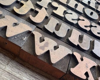 Op de afbeelding: Een verzameling vintage houten letterpress-drukblokken. De blokken zijn gerangschikt in rijen en kolommen en tonen verschillende hoofdletters. Het hout heeft een verweerde uitstraling met kleurvariaties, variërend van licht- tot donkerbruin.