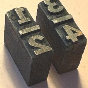 May include: Two vintage letterpress printing blocks. One block features the letter "I" and the letter "S". The other block features the numbers "3" and "4". The blocks are dark gray with silver-colored metal letters and numbers.