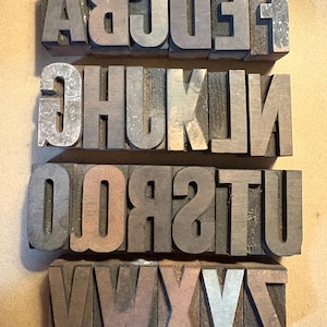 May include: Four rows of vintage wooden letterpress printing blocks. The letters are in various shades of gray and brown, with some showing wear. The letters are arranged in a horizontal format, with each row displaying a different set of letters.