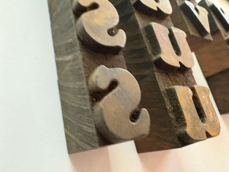 May include: Close-up of weathered, metallic letters, including the letter "S" and "U", mounted on a wooden block. The letters have a rustic, aged appearance, suggesting a vintage or industrial aesthetic. The wooden block has a dark, textured surface.