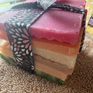 May include: A stack of colorful handmade soaps tied with a patterned ribbon. The soaps are layered in shades of pink, orange, white, and green. The ribbon is black with a floral design. The soaps are on a textured, tan surface.