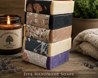 May include: A vertical stack of five rectangular bars of soap, each wrapped in a unique paper band featuring botanical and floral patterns. The soap bars vary in color, including cream, black, light brown, off-white, and purple, arranged neatly.