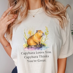 May include: A white t-shirt with a watercolor illustration of a capybara sitting on a rock surrounded by green plants. The text "Capybara Loves You. Capybara Thinks You're Great." is printed below the illustration.
